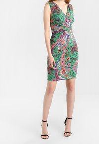 Grön paisleymönstrad klänning med omslagsdesign och V-ringning. Har livliga mönster och en figurnära silhuett, som kombineras med svarta stilettklackar.