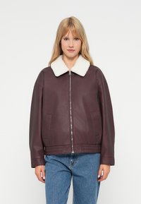 Giacca in pelle bordeaux scuro con colletto in shearling bianco, chiusura con zip, vestibilità ampia e tasche laterali, indossata sopra jeans blu.