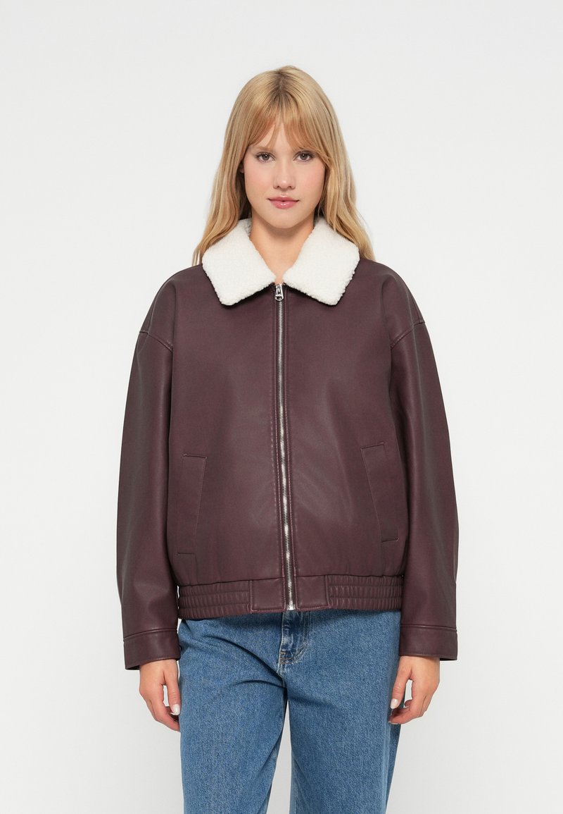 Giacca in pelle bordeaux scuro con colletto in shearling bianco, chiusura con zip, vestibilità ampia e tasche laterali, indossata sopra jeans blu.