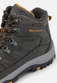 Scarponcino da trekking grigio con materiale idrorepellente, tomaia testurizzata, accenti neri e arancioni, occhielli rinforzati e suola in gomma robusta.