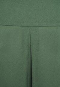 Tessuto verde con una superficie liscia, caratterizzato da un dettaglio di cucitura verticale. Il materiale appare leggero e scorre delicatamente.