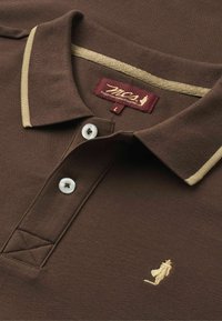 Polo marron avec liseré beige au col, deux boutons blancs, logo brodé représentant un homme qui marche et étiquette bordeaux près de l’encolure.