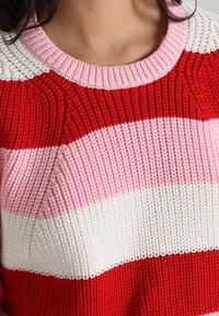Pull en maille avec rayures horizontales rouge, rose, rose clair et blanc. Matière texturée avec un col arrondi.