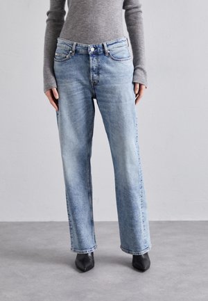 Personne portant un jean large bleu clair, un haut gris côtelé à manches longues et des chaussures noires à bouts pointus, debout sur un sol gris.