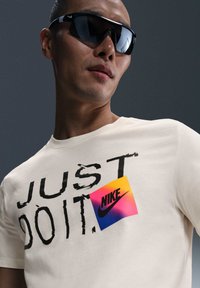 Camiseta de algodón crema con texto impreso en negro "JUST DO IT" y un parche colorido del logo de Nike. Modelo usando gafas de sol negras frente a un fondo gris.