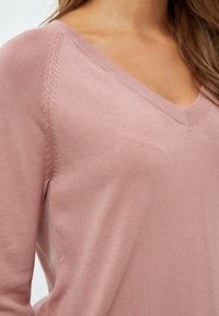 Pulover z V-izrezom v mehki rožnati tkanini, z rebrastim ovratnikom in dekorativnim šivanjem vzdolž rokavov. Gladka tekstura in prilegajoča zasnova.