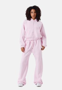 Sudadera rosa claro y pantalones a juego, ambos en tejido suave con un corte relajado, con cierre de cremallera y bolsillos frontales.