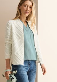 Veste bomber blanche matelassée avec texture en zigzag, assortie à un blouse bleu clair en v-neck et un jean bleu. Tenant un sac vert avec une sangle.