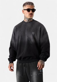 Schwarz Oversized-Sweatshirt mit geripptem Kragen und Bündchen, ausgestattet mit einem verblassten Design und einem kleinen Logo auf der linken Brust. Matte Textur.
