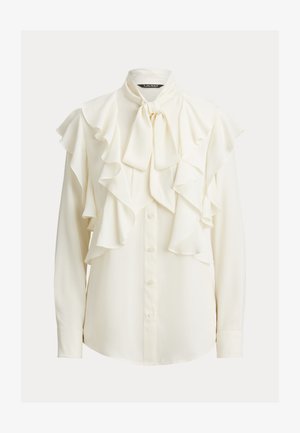 Blusa bianca con accenti arricciati lungo il petto e le maniche, un fiocco legato al colletto e dettagli con bottoni sulla parte anteriore. Tessuto morbido.