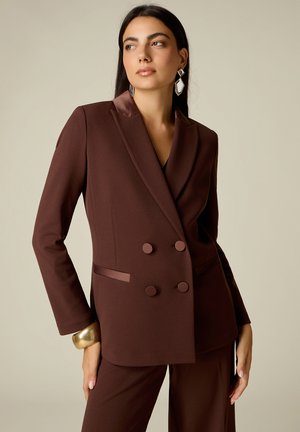 Kvinde med langt mørkt hår iført en brun, dobbeltradet blazer og matchende bukser, med geometriske øreringe og et stort gyldent armbånd.