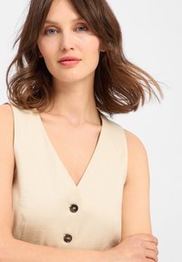 Beige Leinen-Top mit tiefem V-Ausschnitt, ausgestattet mit zwei dunklen Knöpfen vorne. Sanfte Textur, ärmelloses Design und lockerer Schnitt.