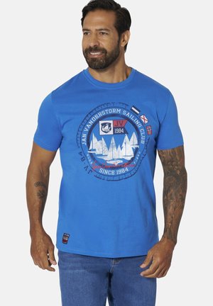 Man with beard wearing blue short-sleeve sailing club t-shirt with white sailboat graphic and nautical flags, paired with blue jeans.
