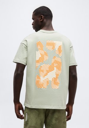 Mężczyzna ubrany w jasnozieloną koszulkę z dużym abstrakcyjnym pomarańczowym paskowanym wzorem na plecach oraz zielone krótkie spodenki moro z napisem "TERREX".