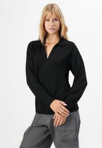 Mulher com cabelo loiro, vestindo um suéter preto de decote em V e calças cargo cinzas, de pé com os braços cruzados à frente contra um fundo branco.