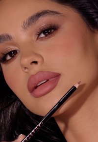 Konturówka do ust z ostrym końcem trzymana w pobliżu pełniejszych warg, w odcieniu nude. Widoczna gładka tekstura i eleganckie czarne opakowanie.