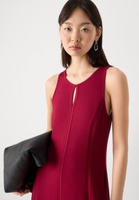 Ribattu, ilman hihoja oleva mekko syvässä punaisessa, jossa on avainaukko kaula-aukko. Malli pitää mustaa nahkakosketinta kädessään. Yksinkertainen, tyylikäs muotoilu.