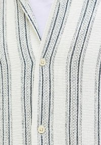 Tissu blanc texturé avec des rayures verticales cousues en noir et gris, comprenant deux boutons crème sur un vêtement à encolure en V.