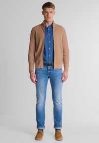 Beżowy sweter z dzianiny z wysokim kołnierzem noszony na niebieskiej koszuli z dżinsu, w połączeniu z jasnoniebieskimi dżinsami i beżowymi butami. Prosty krój z dopasowaną sylwetką.