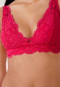 Bralette in pizzo rosa acceso con scollo a V, motivi floreali, bordi smerlati e dettaglio a chiave al centro. Tessuto morbido e testurizzato.