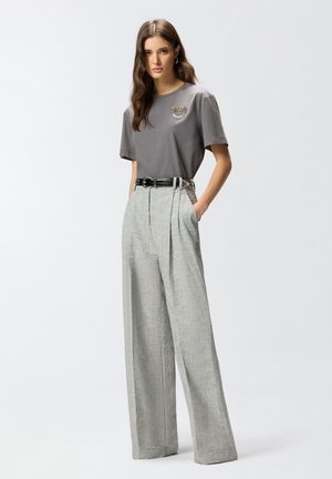 Femme aux longs cheveux bruns portant un t-shirt gris avec un détail brodé, rentré dans un pantalon gris taille haute à jambes larges avec une ceinture noire.