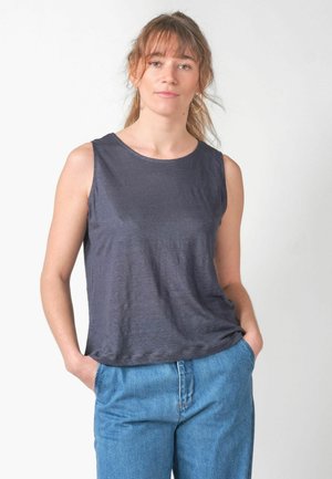 Mladá žena so svetlohnedými vlasmi zviazanými do copu, oblečená v tmavosivom tielku bez rukávov a modrých džínsoch, stojaca s rukami v kapsách.