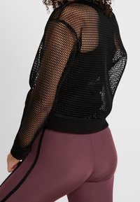 Czarny siatkowy top z długim rękawem o luźnym kroju, z prążkowanymi czarnymi mankietami i jednolitym czarnym dołem. Noszony na czarnym dopasowanym topie na ramiączkach.