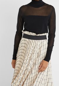 Top lungo a maniche trasparenti nere con collo alto, abbinato a una gonna plissettata a quadri beige con elastico nero in vita.