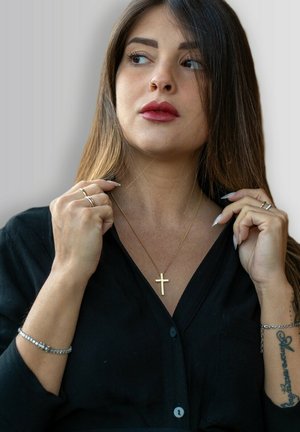 Goldenes Kreuz-Halsband mit einer dünnen Kette, getragen über einem schwarzen Shirt. Das Modell hat lange braune Haare und trägt silberne Ringe und ein Armband.