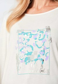Weißes Langarmhemd mit einem hellblauen, lila und silbernen Grafikdesign, das den Text "Das Leben ist schön" und glänzende Akzente enthält.