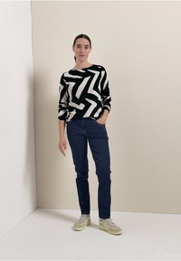 Suéter de punto con estampado de zigzag en blanco y negro, combinado con jeans de azul oscuro y zapatillas claras. El modelo se encuentra de pie contra un fondo neutro.