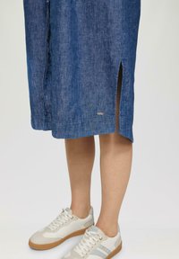 Denimkleid mit seitlichem Schlitz, versehen mit einem blauen strukturierten Muster. Kombiniert mit weißen Sneakers, cremefarbenen Akzenten und einer Gummisohle.