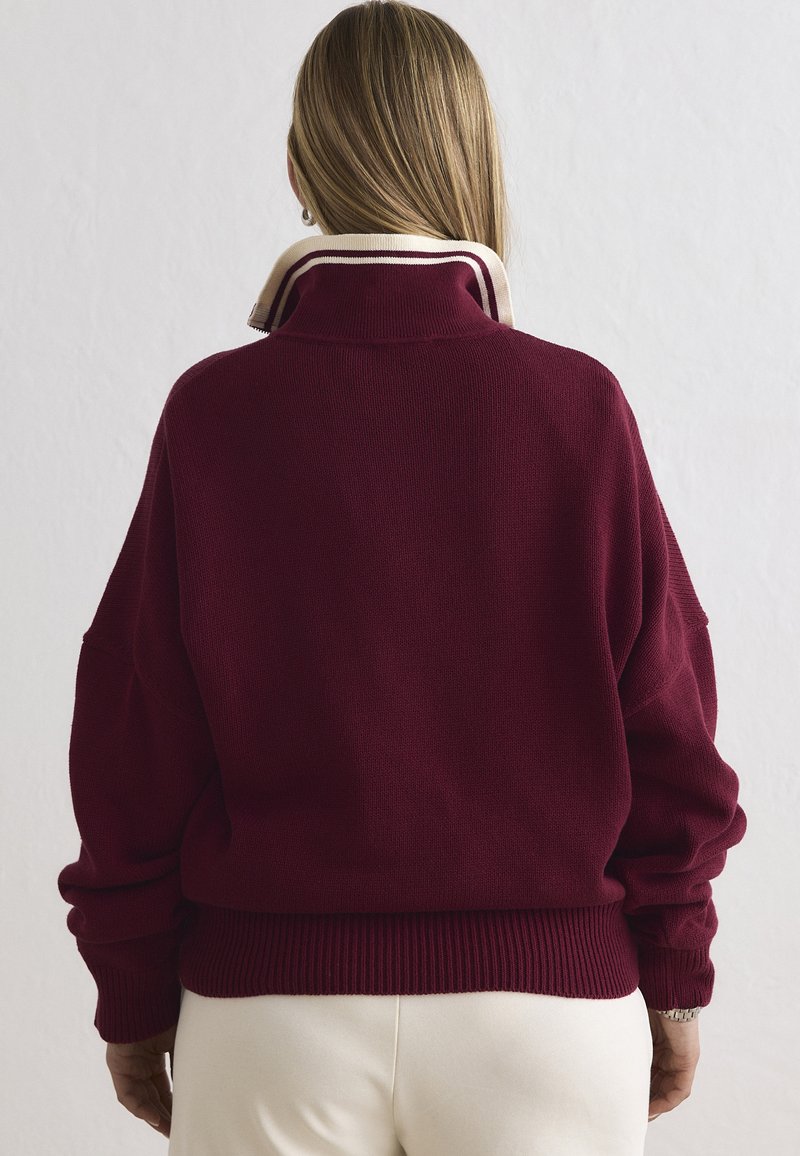 Femme aux longs cheveux blonds portant un pull bordeaux à col montant et un pantalon couleur crème, vue de dos.