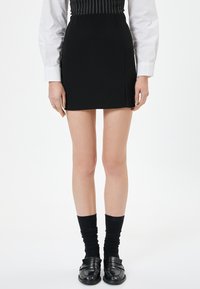 Mini gonna nera con taglio dritto e spacco laterale, abbinata a calze nere con texture e mocassini lucidi, indossata con una camicia bianca a maniche lunghe.