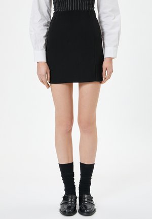 Zwarte minirok met een rechte snit en zijslit, gecombineerd met zwarte textuur sokken en glanzende loafers, gedragen met een wit shirt met lange mouwen.