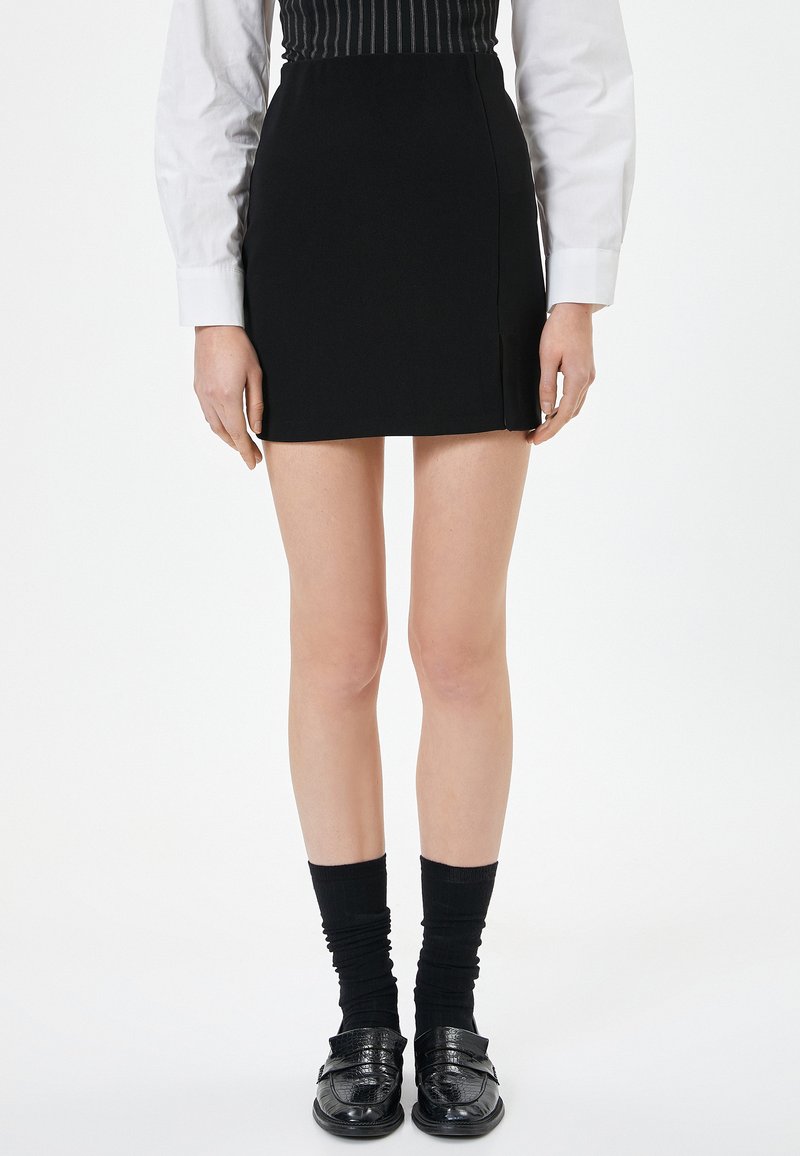 Mini gonna nera con taglio dritto e spacco laterale, abbinata a calze nere con texture e mocassini lucidi, indossata con una camicia bianca a maniche lunghe.