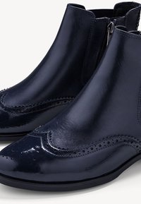 Bottes chevilles en cuir bleu marine brillant avec des détails brogue, une texture lisse et une fermeture éclair latérale pour un enfilage facile.