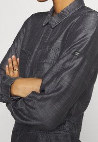 Camisa de color gris oscuro con textura, con cuello, dos bolsillos en el pecho y mangas largas. Presenta una pequeña etiqueta en la manga.