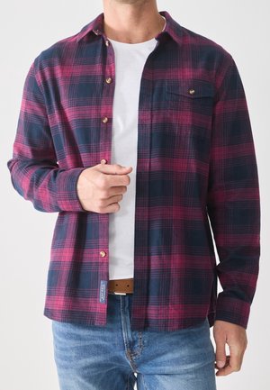 Mand iført åben rød og mørkeblå ternet skjorte over hvid t-shirt og blå jeans, justerer skjorten med venstre hånd.