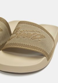 Ciabatte color cuoio con cinturino intrecciato testurizzato e suola liscia color crema. Il cinturino presenta un motivo sottile e un dettaglio di branding.