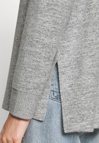Pull en maille gris avec des poignets côtelés, des fentes sur les côtés et une texture douce ; porté sur un denim bleu, mettant en valeur des styles et des couleurs contrastants.