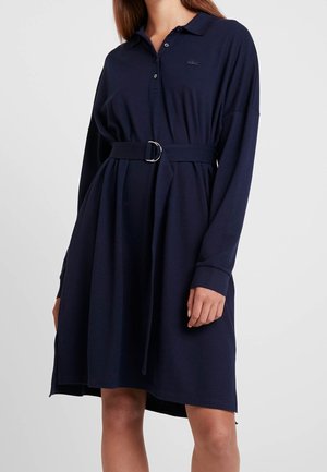 Kobieta w granatowej, długorękawowej sukience polo z paskiem, guzikami i logo na piersi, stojąca na jednolitym tle.