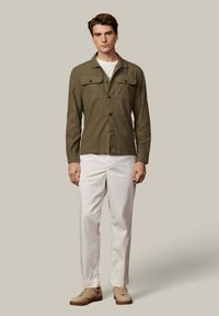 Chaqueta abotonada verde oliva con dos bolsillos en el pecho, usada sobre una camiseta clara, combinada con pantalones blancos y zapatos beige. Textura de tela suave.