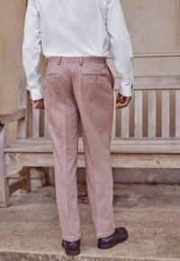 Helle rosa Hosen mit gewebter Textur, zwei Gesäßtaschen und goldfarbenen Knopfdetails, kombiniert mit einem weißen Langarmshirt und dunklen Schuhen.