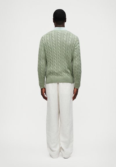 Ανοιχτό πράσινο πουλόβερ με πλέξη καλωδίου και ούλα περιθώρια, φορεμένο πάνω από λευκά, φαρδιά λινά παντελόνια. Η πλάτη διαθέτει περίπλοκους υφασμάτινους λεπτομέρειες.