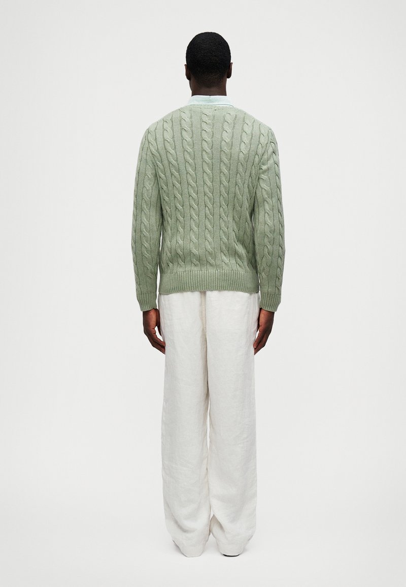 Suéter de punto trenzado verde claro con dobladillo acanalado, usado sobre pantalones de lino blancos y holgados. La parte posterior presenta un intrincado detalle texturizado.