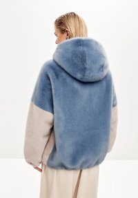 Cappotto in pelliccia blu e beige con cappuccio, caratterizzato da una vestibilità ampia e una texture morbida. Il design evidenzia blocchi di colore a contrasto sulle maniche.