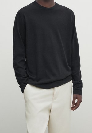 Mann in einem schwarzen langärmligen Pullover, der leicht in weiße Hose gesteckt ist, steht mit einer Hand in der Tasche vor einem schlichten Hintergrund.