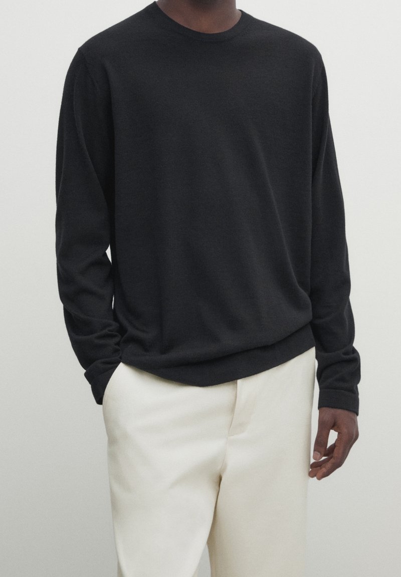 Homme portant un pull noir à manches longues légèrement rentré dans un pantalon blanc, se tenant avec une main dans la poche devant un fond uni.