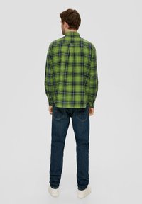Green plaid button-up shirt with long sleeves, featuring dark blue lines and a back pleat, paired with dark blue jeans and white sneakers.